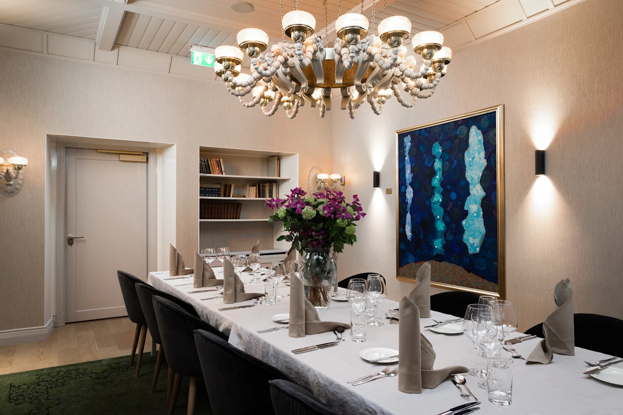 A dining table is elegantly set with folded napkins and wine glasses, surrounded by black chairs. A chandelier hangs overhead in a modern room with artwork on the wall.