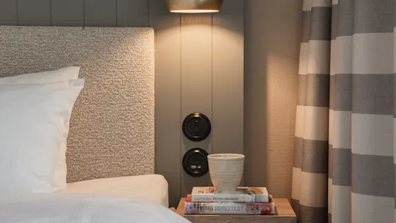 A cozy bedside scene in a hotel room with a warm lamp, books, and a cup on a nightstand next to a comfortable bed.