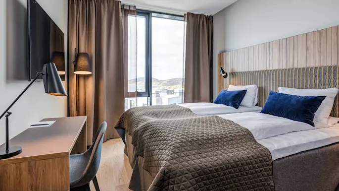 A neatly made bed with blue pillows and brown blanket faces a window with curtains, accompanied by a wooden desk, chair, and TV in a modern room.