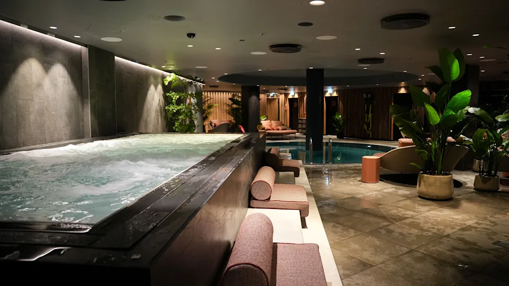 A luxurious indoor spa area with a large bubbling hot tub, lounge chairs, and a serene swimming pool in the background.
