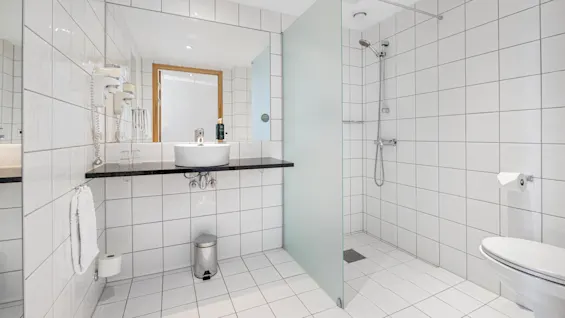 A clean, modern bathroom with a sink, mirror, shower, and toilet in a tiled, well-lit space.