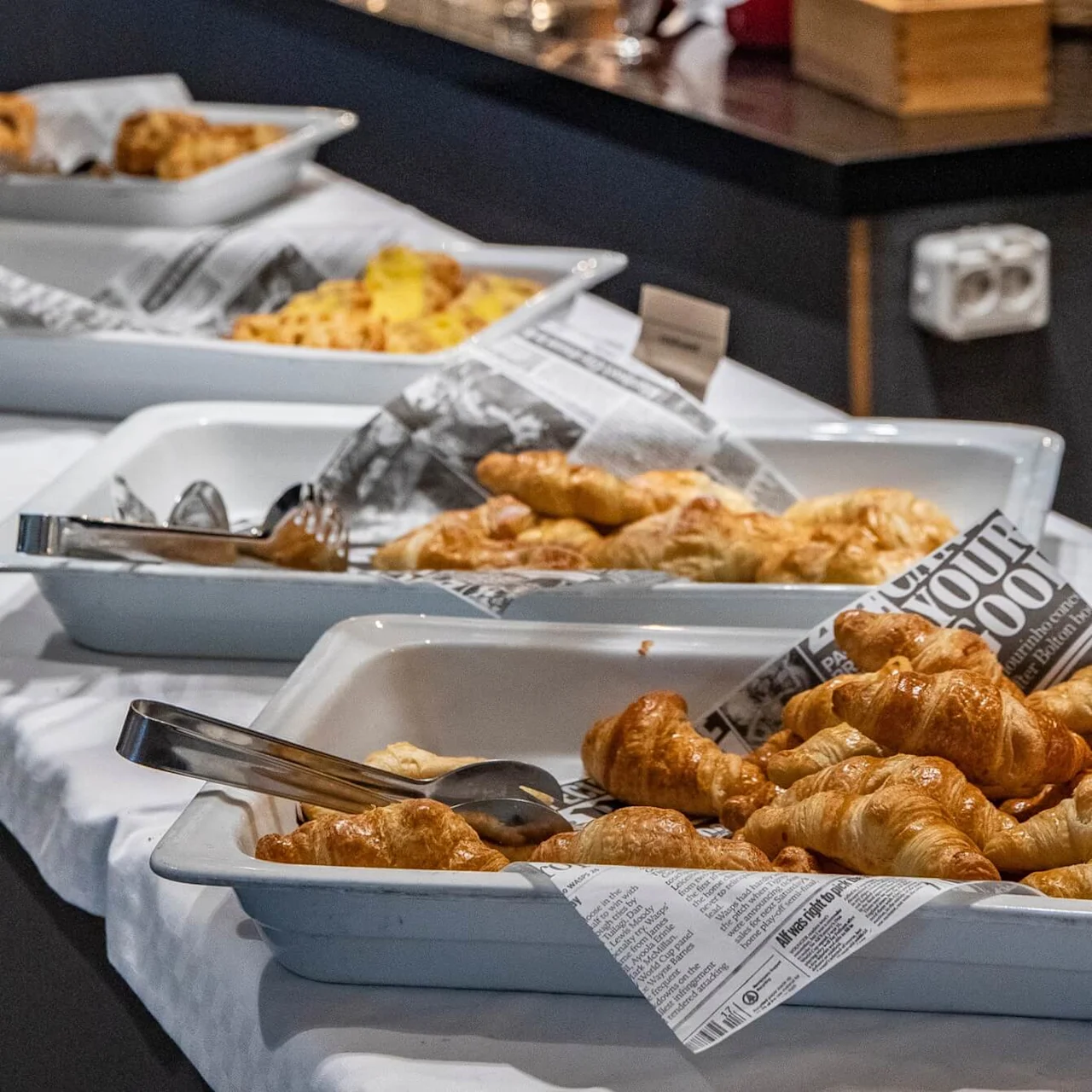Croissanter ligger i hvite brett belagt med avismønsterpapir; metalltang hviler i brettene. Flere brett med bakverk er justert på en hvit duk i en buffésetting.
