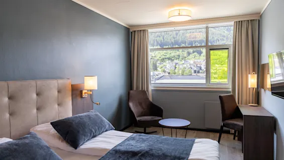 En snyggt bäddad säng med mörka kuddar och överkast står i ett hotellrum upplyst av lampor. Ett fönster visar en scenisk vy av kullar och träd.