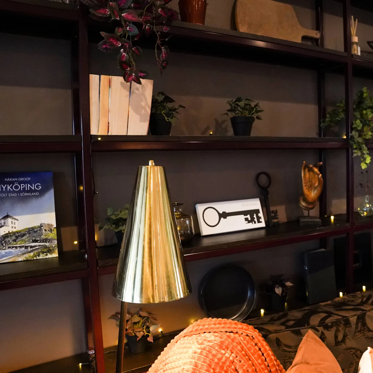 A bookshelf is decorated with plants, books, and various objects in a cozy room with warm lighting.