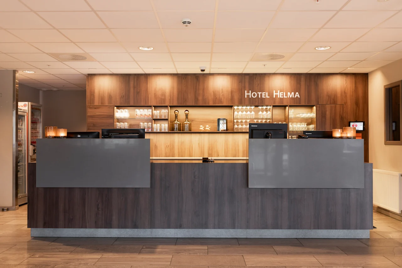 Et moderne hotelreceptionsområde med en trædisk og oplyste hylder. Teksten HOTEL HELMA vises ovenfor, klar til gæster.