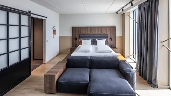 Et moderne soverom har en stor seng med hvitt sengetøy. En mørk sofa står ved fotenden av sengen. En skyvedør og gardiner forbedrer den stilige, minimalistiske innredningen.