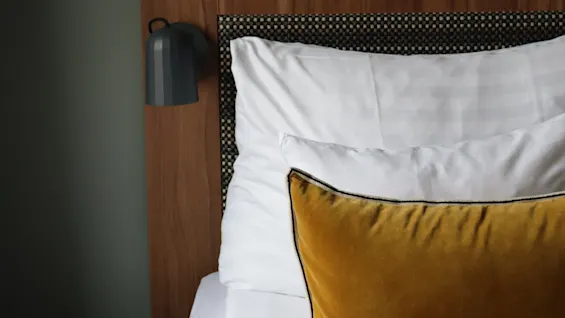 Bed adorned with a mustard yellow pillow rests under a wooden headboard. A wall-mounted light fixture and a small black side table enhance the minimalist bedroom design.