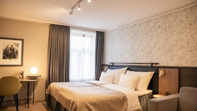 En pent redt seng, med beige sengetøy, står i et fredelig soverom. I nærheten er det et lite skrivebord med lampe, stoler, og mønstret tapet som fremhever den koselige atmosfæren.