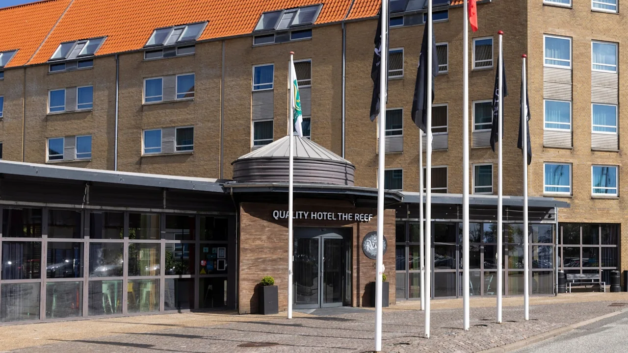Hotellets indgang med skiltet "QUALITY HOTEL THE REEF" er omgivet af høje flagstænger. Bygningen har flere vinduer, et orange tegltag og en klar blå himmel i baggrunden.