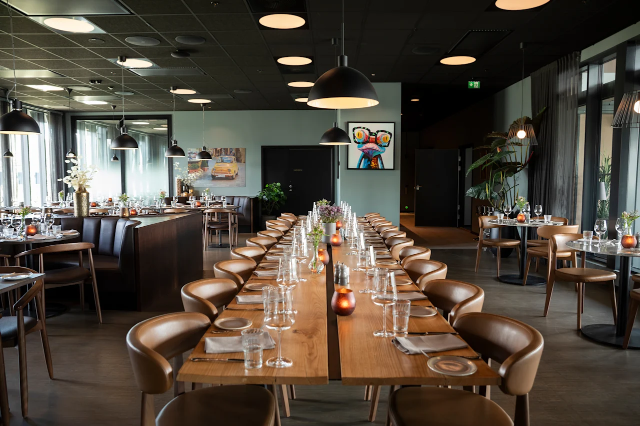 Et moderne restaurantinteriør med et langt spisebord dekket for en stor samling, omgitt av mindre bord.