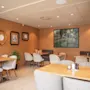 Tables and chairs arranged in a cozy café setting, surrounded by warm, earthy-colored walls adorned with framed art. Ambient lighting creates a relaxed atmosphere.