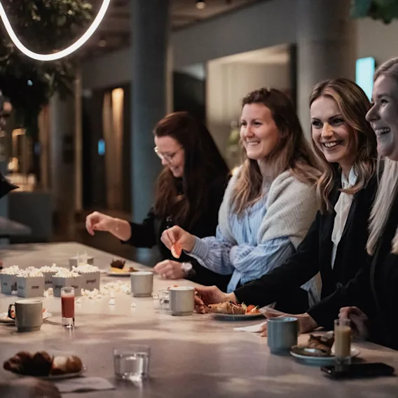 Fyra kvinnor ler och umgås vid ett bord med drycker och snacks i ett svagt upplyst rum.