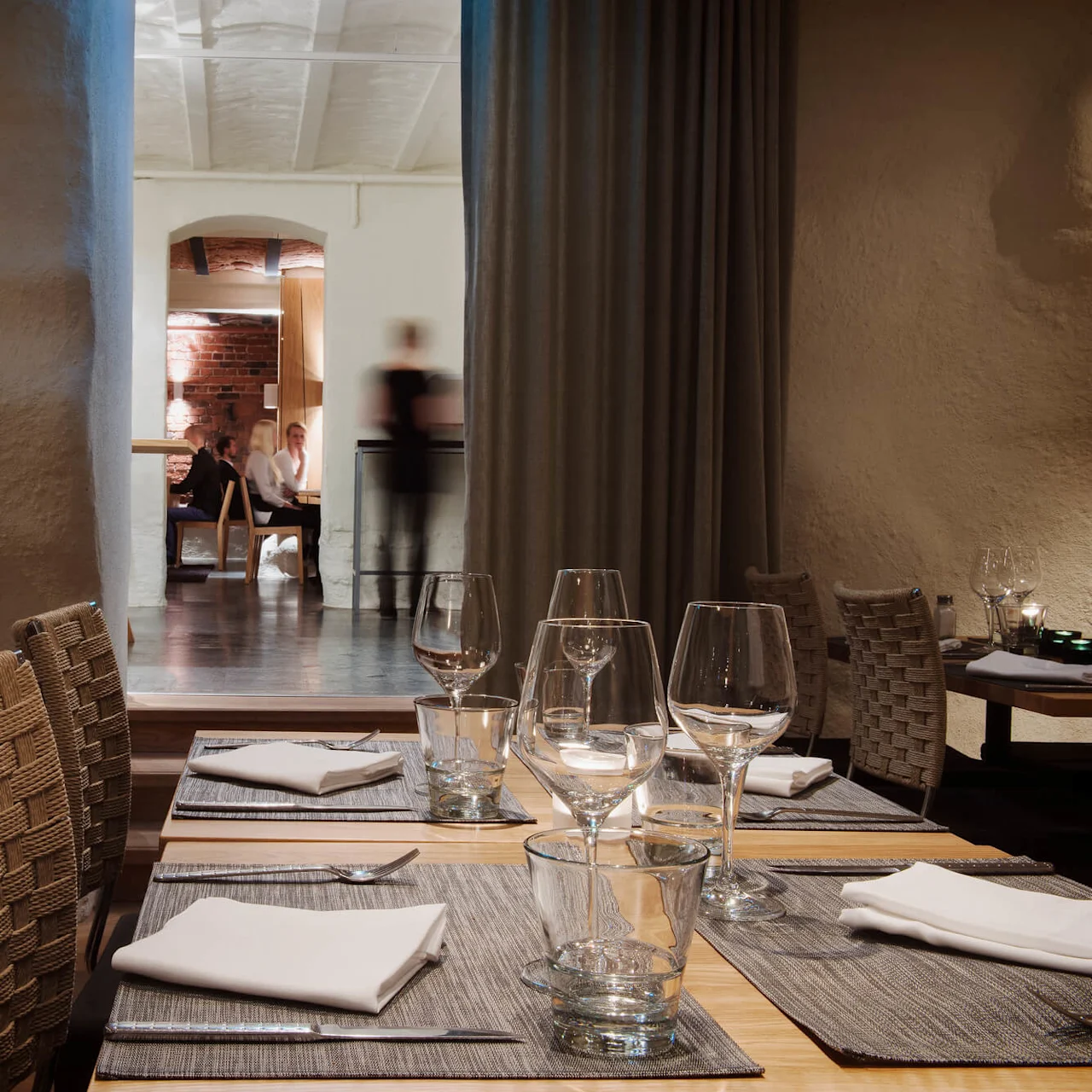 Et spisebord med pænt arrangerede glas og servietter i en elegant restaurant. Baggrunden viser slørede personer i samtale nær en murstensvæg, hvilket giver en hyggelig stemning.