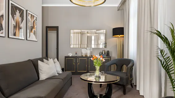 En stilfuld hotelsuite stue med grå sofa, sofabord med blomster, lænestol og spejlskab.