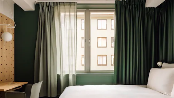 A modern hotel room at Hobo Oslo with a bed, desk, and window with dark green curtains.
