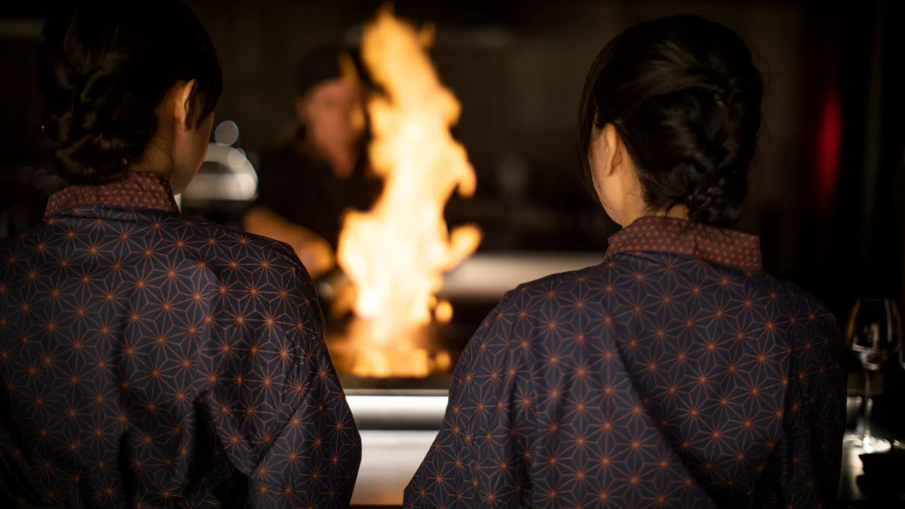 Two people in patterned uniforms watch a chef cooking with flames, in a restaurant kitchen.