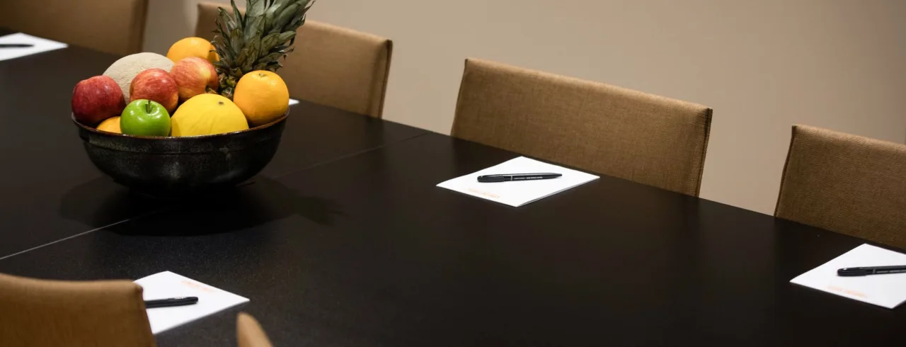 A bowl of assorted fruits on a black conference table with papers and pens, surrounded by chairs.