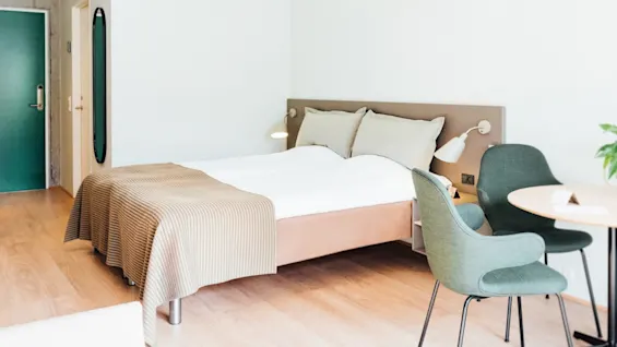 En pænt redt seng med hvid dyne og beige tæppe står ved siden af ​​et lille rundt bord med to grønne stole i et moderne, minimalistisk rum med trægulv.