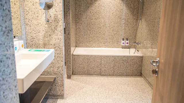 Kylpyhuone, jossa valkoinen pesuallas, kylpyamme suihkupäällä ja hygieniatuotteita; epäkeskosten kiviseinien ja lattian keskellä, luoden tyylikkään, modernin ilmeen. Pullot sijoiteltu ammeen reunalle.