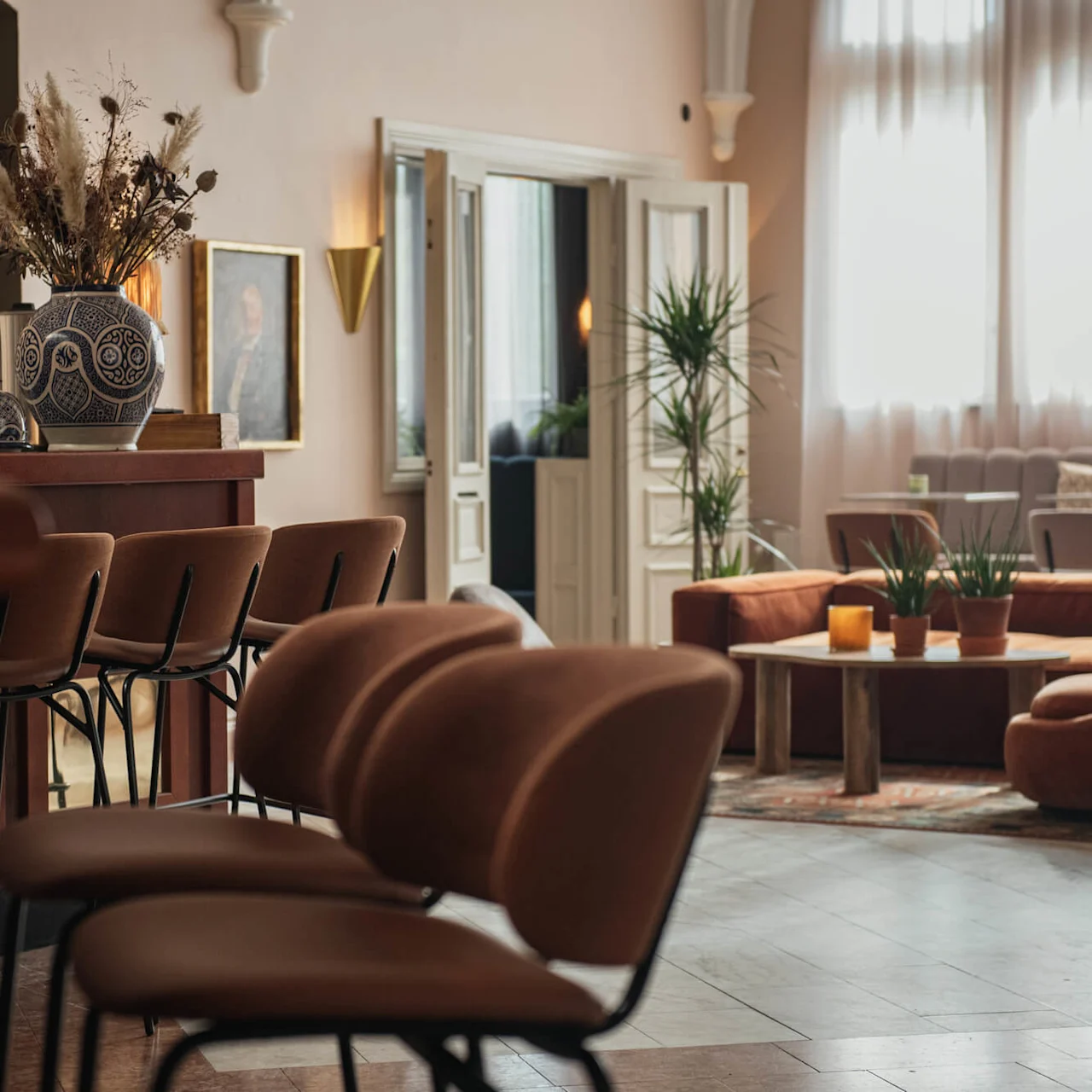 Chairs line a wooden bar adorned with decorative vases and dried plants; the room features soft lighting, a painting on the wall, and a lounge area with sofas and potted plants.