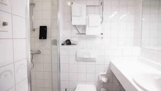 Hvidt flisebelagt badeværelse; vægmonteret håndklædeholder med hvide foldede håndklæder over et toilet. Bruser med glasdør til venstre, håndvask med bordplade til højre. Hårtørrer på væggen.