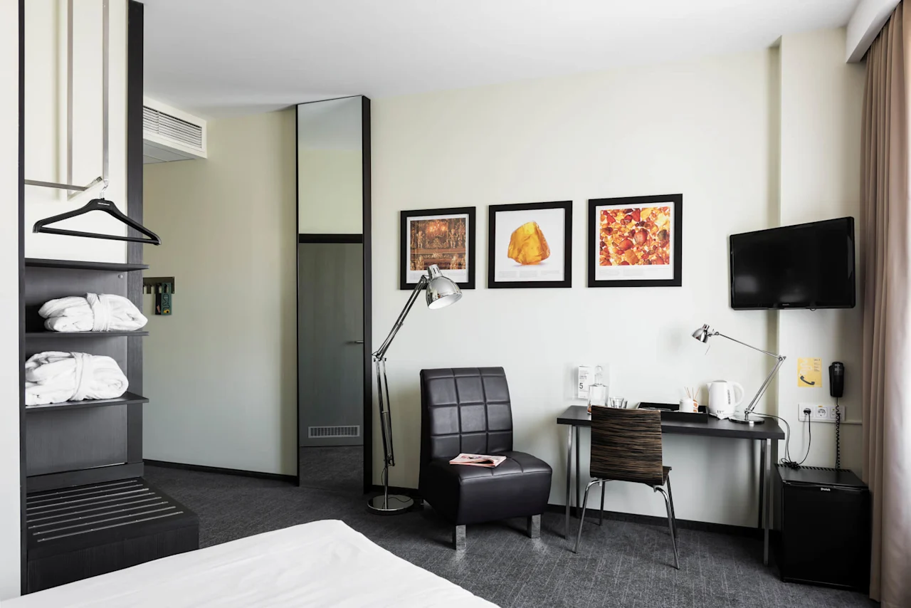 A hotel room features a modern chair beside a lamp, near a wall with three framed artworks. A desk holds various items, opposite a small television on the wall.