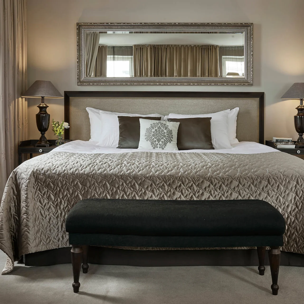 A large bed with pillows and a textured comforter is centered in a bedroom. Two lamps on nightstands flank the bed, with a large mirror above. Light streams through curtains.