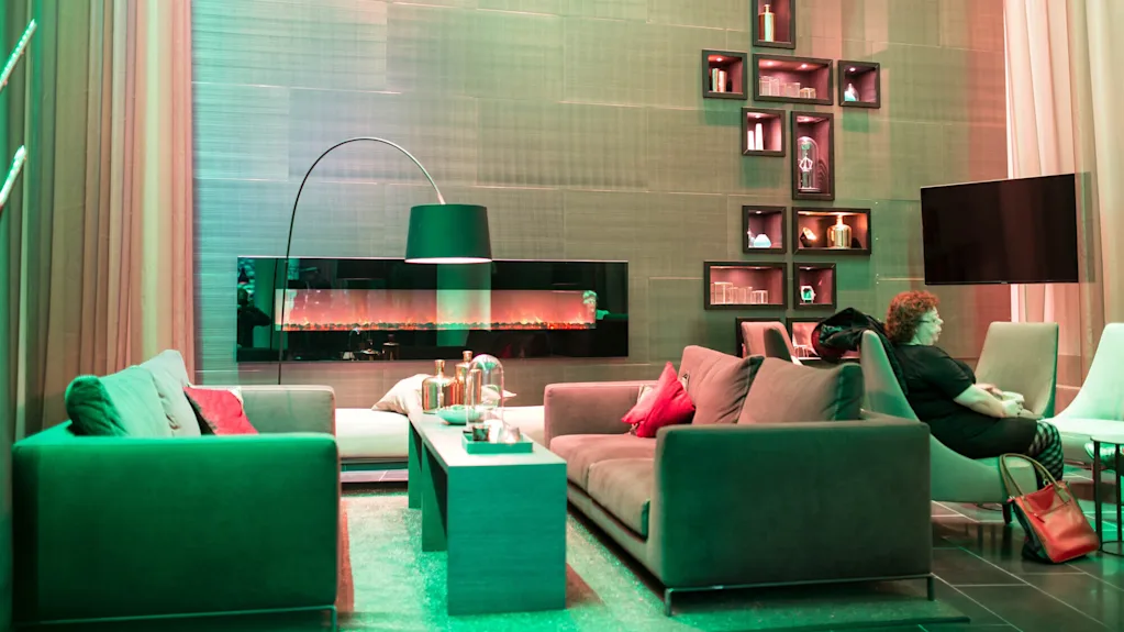 En modern lounge har två grå soffor mittemot varandra med ett bord emellan, under en stor vägglampa. En dekorativ vägg och elektrisk eldstad ger stämning.