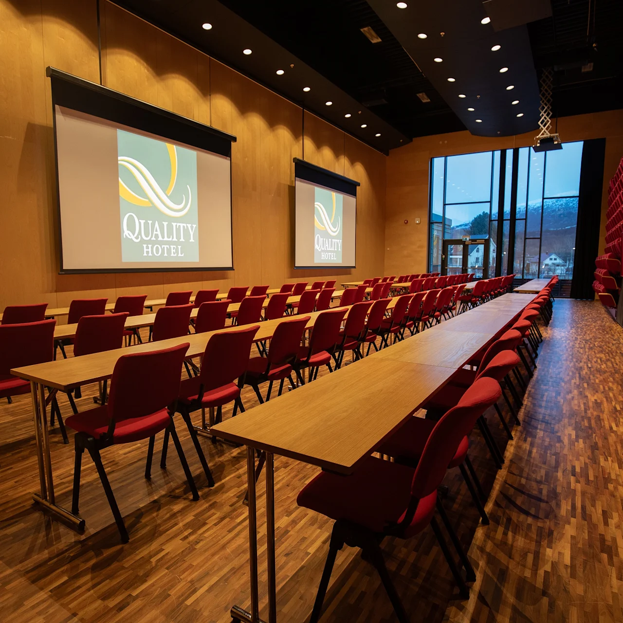 Rader av røde stoler og trebord vender mot to projeksjonsskjermene som viser "QUALITY HOTEL" i et stort, moderne konferanserom med trepanel og et høyt vindu med utsikt over fjellene.