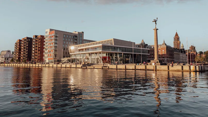 Modern waterfront buildings reflect on calm water, surrounded by traditional architecture and a statue on a tall column. The scene is set during sunset with soft, warm lighting.