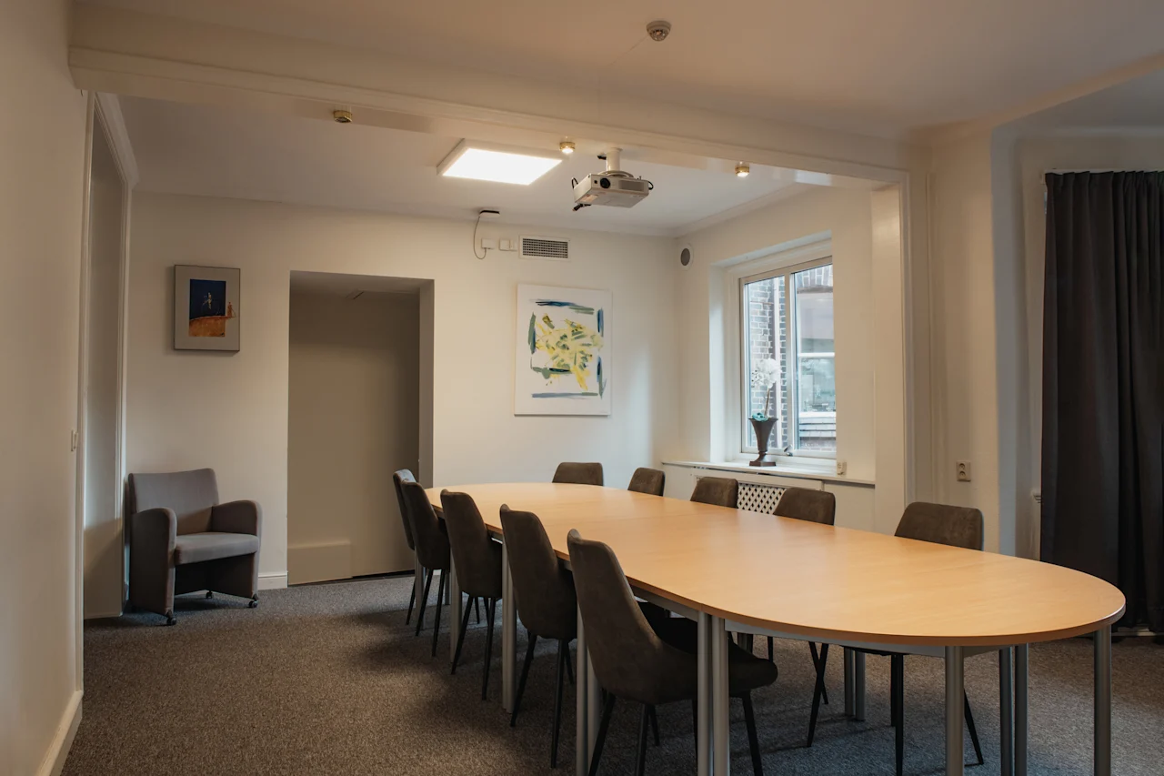 Et veloplyst konferencelokale med et stort ovalt bord og stole, klar til et møde eller en præsentation.