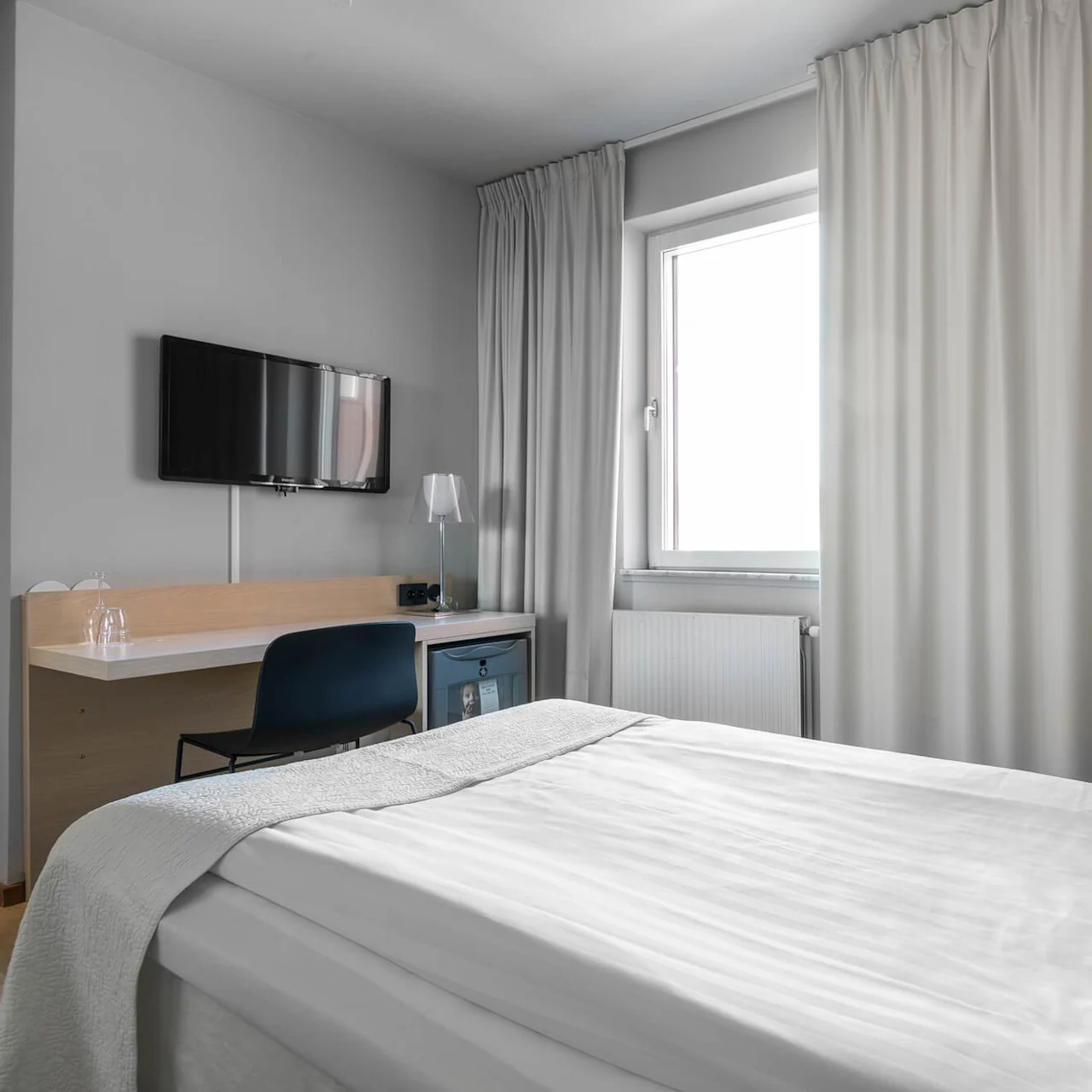 En prydlig hotellrum med en snyggt bäddad säng. En väggmonterad TV och ett skrivbord med en stol är placerade nära ett fönster täckt av ljusa gardiner.