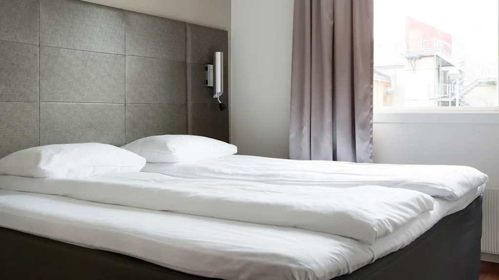 A neatly made bed with white linens and pillows sits in a modern room. Light gray curtains frame a window, revealing an outdoor staircase and building.