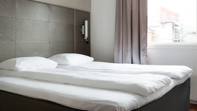 Sängyssä on valkoiset vuodevaatteet ja tyynyt modernissa huoneessa. Vaaleanharmaat verhot kehystävät ikkunan, josta näkyy ulkoportaat ja rakennus.