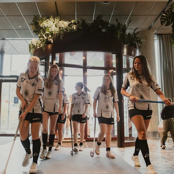 En grupp unga idrottare i uniformer med innebandyklubbor går genom en hotellobby. Mattan lyder Dina första steg till en magisk vistelse.