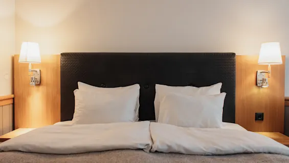 A comfortable hotel bed with white linens and warm lighting from wall-mounted lamps, ready for a restful stay.