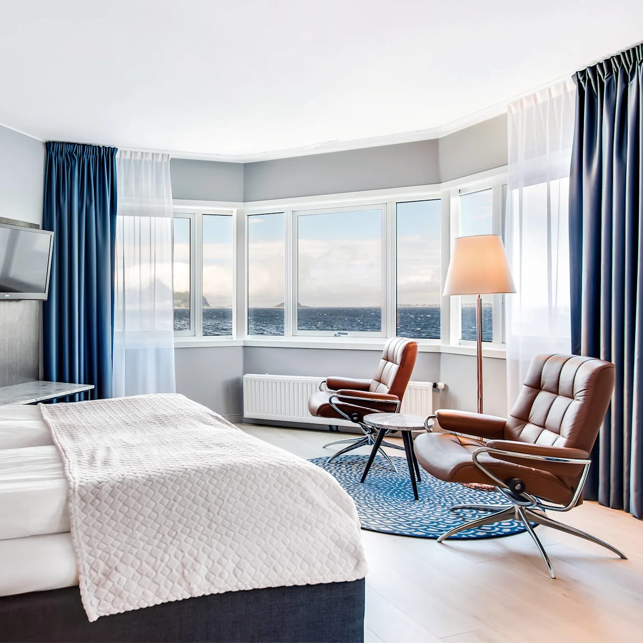 Et pænt indrettet hotelværelse med en seng med mønstrede puder, to brune stole vendt mod en udsigt over havet gennem store vinduer, blå gardiner og et tv på væggen.