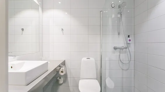 Et hvidt toilet står mellem en rektangulær vask og et bruseområde med glasvæg i et flisebelagt badeværelse med minimalistisk indretning.