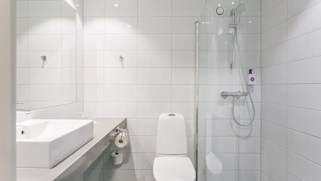 En vit toalett står mellan en rektangulär handfat och en dusch med glasvägg i ett kaklat badrum med minimalistisk inredning.