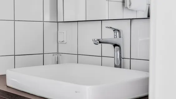 Modern sink sits atop a wooden counter, featuring a sleek chrome faucet. Surrounded by white tiled walls, a towel hangs nearby, creating a clean, minimalist bathroom setting.