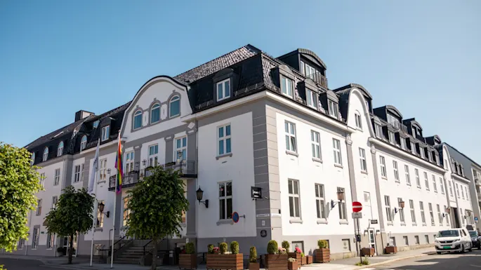 A white and grey building with a dark roof, trees, and a rainbow flag. A street with a crosswalk, a stop sign, and text JAREN.