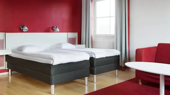 Two twin beds with white sheets rest in a hotel room with red walls and curtains. A red sofa and round white table are nearby. Natural light streams through a window.