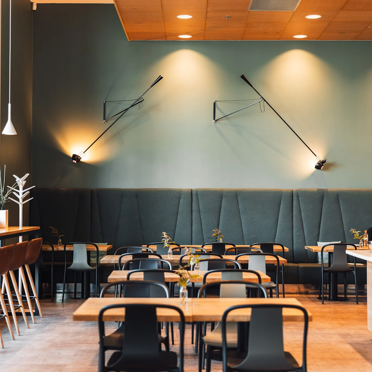 Tables and chairs are neatly arranged in a modern dining area with minimalistic wall lamps, green padded wall panels, and a wooden ceiling, creating a contemporary ambiance.