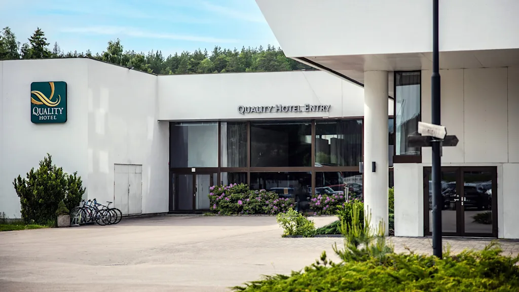En moderne hotellinngang med store glassdører, omgitt av hvite vegger, har skiltene "QUALITY HOTEL" og "QUALITY HOTEL ENTRY". Sykler og grøntområder forbedrer settingen.
