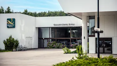 En moderne hotellinngang med store glassdører, omgitt av hvite vegger, har skiltene "QUALITY HOTEL" og "QUALITY HOTEL ENTRY". Sykler og grøntområder forbedrer settingen.