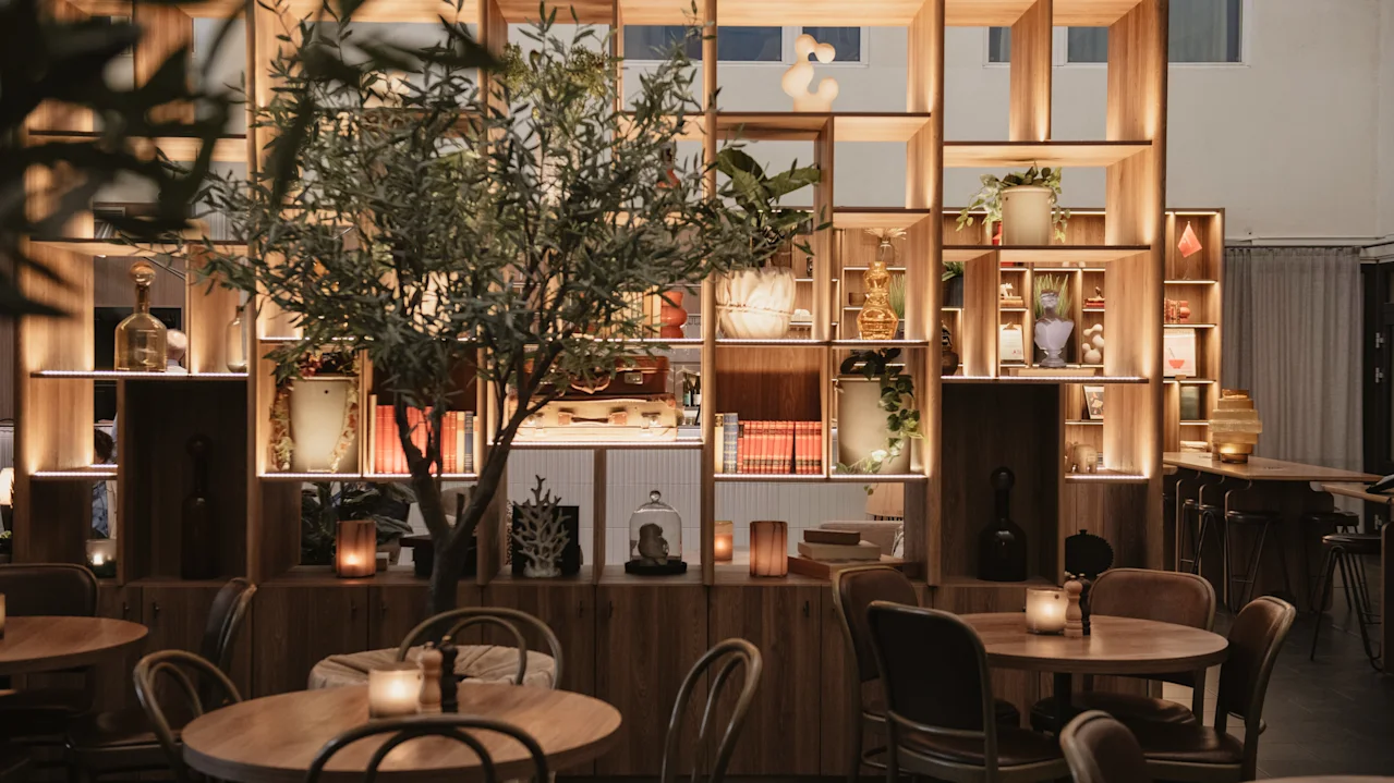 Bord med stearinlys er arrangerede foran en træhylde, der viser vaser og dekorationer, sat i en varm oplyst, hyggelig café eller restaurant, med et potte træ.