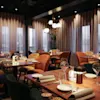 Elegante spisebord er ryddig arrangert, dekket med tallerkener, glass og servietter, under dempet, varm belysning i en moderne restaurant med store vinduer og tynne gardiner.