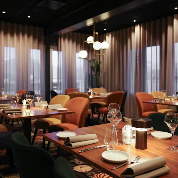 Elegante spisebord er ryddig arrangert, dekket med tallerkener, glass og servietter, under dempet, varm belysning i en moderne restaurant med store vinduer og tynne gardiner.