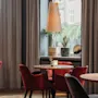 Et varmt og innbydende kafé- eller restaurantinteriør med runde trebord, plysj-røde stoler og planter ved store vinduer.