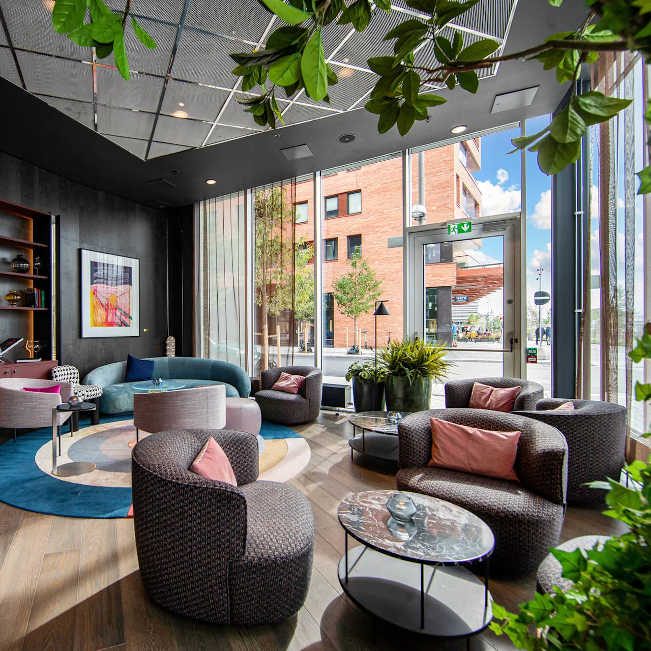 A modern lounge features stylish furniture, including gray chairs and a blue sofa, arranged around tables. Large windows offer street views, and shelves display art and decor.